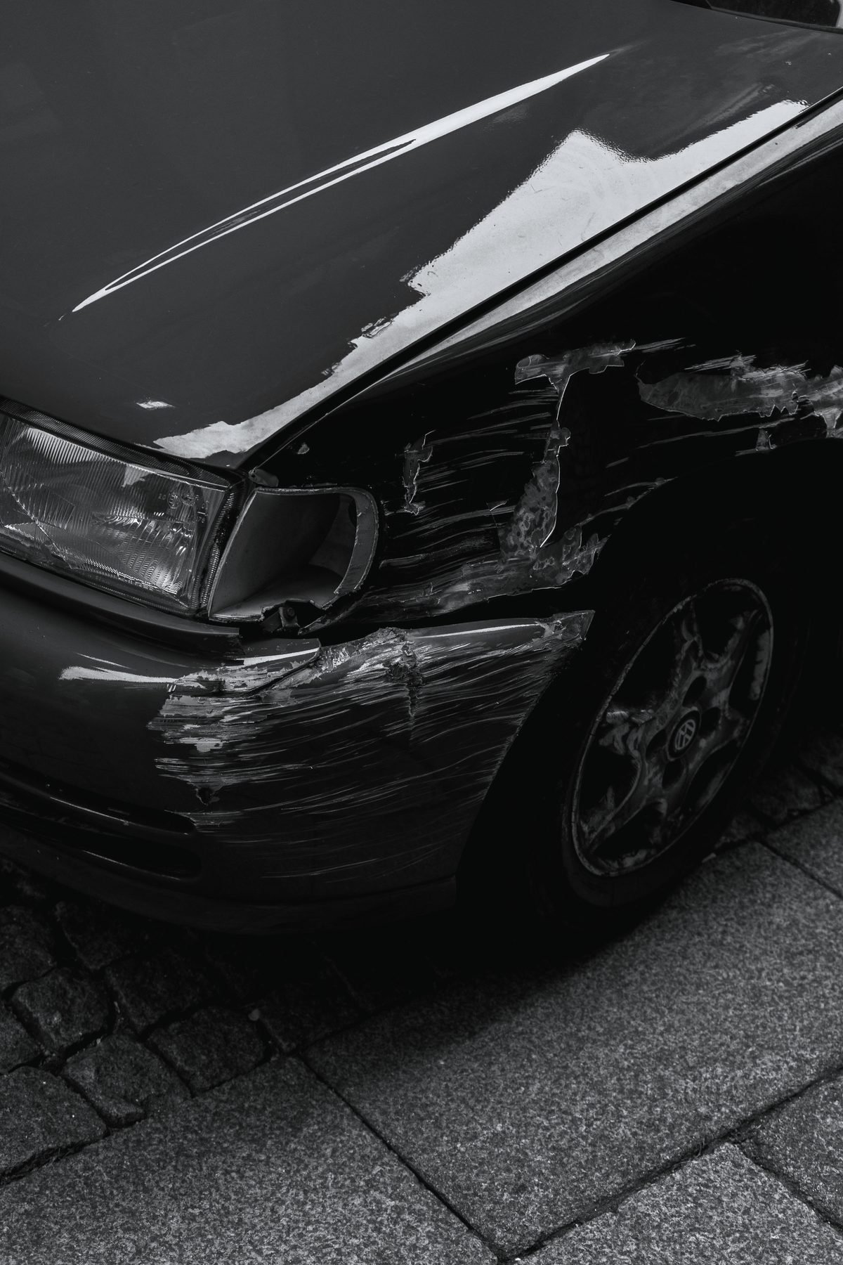 The crumpled front corner of a dark car, headlight and fender pushed in