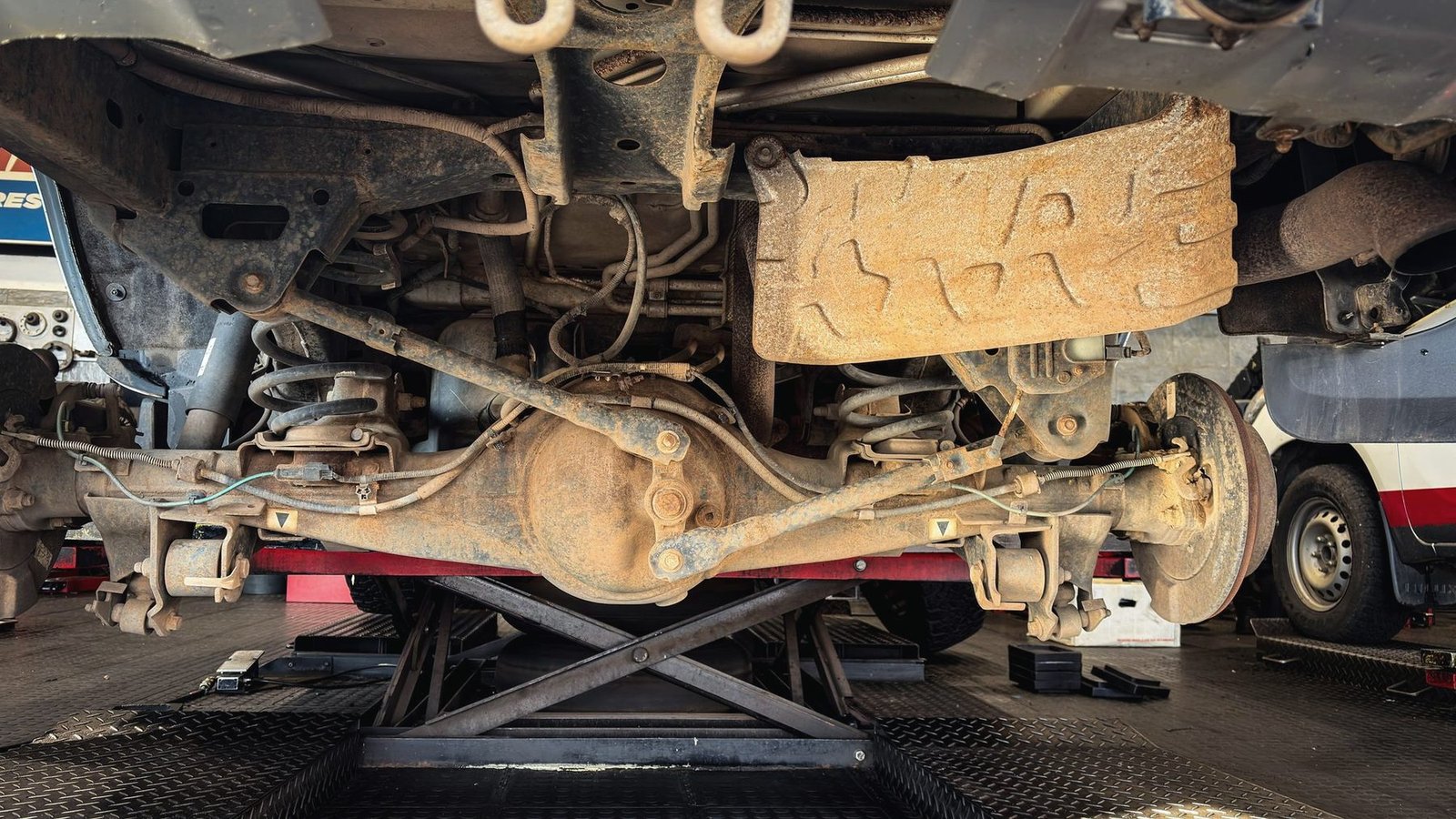 Underside of a vehicle on a lift showing the axle, rails and rust on the floor pan
